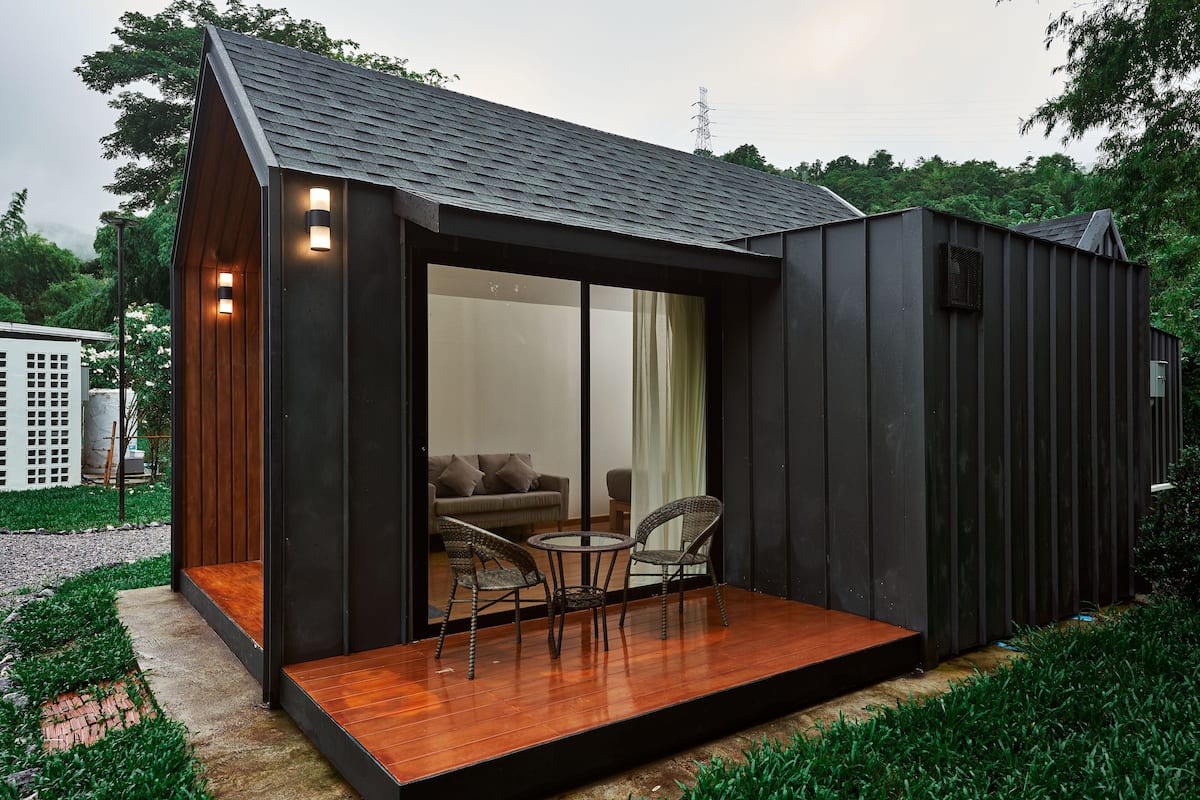 Una casa moderna y compacta con una terraza de madera, una puerta de cristal que da a un interior con sofá y sillones, rodeada de follaje.