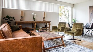 A spacious living room with a wooden coffee table, a sofa, and two armchairs. There is a large window with blinds, a potted plant, and a painting on the wall. - Luxurious Villa in Reykjavik (Reykjavík)