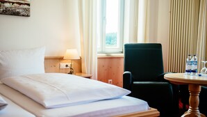 Habitación de hotel con cama, una silla, una mesa, una lámpara y un ventanal con cortinas.