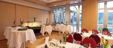 A dining area with multiple tables set for service, featuring white tablecloths, red chairs, and floral centerpieces.