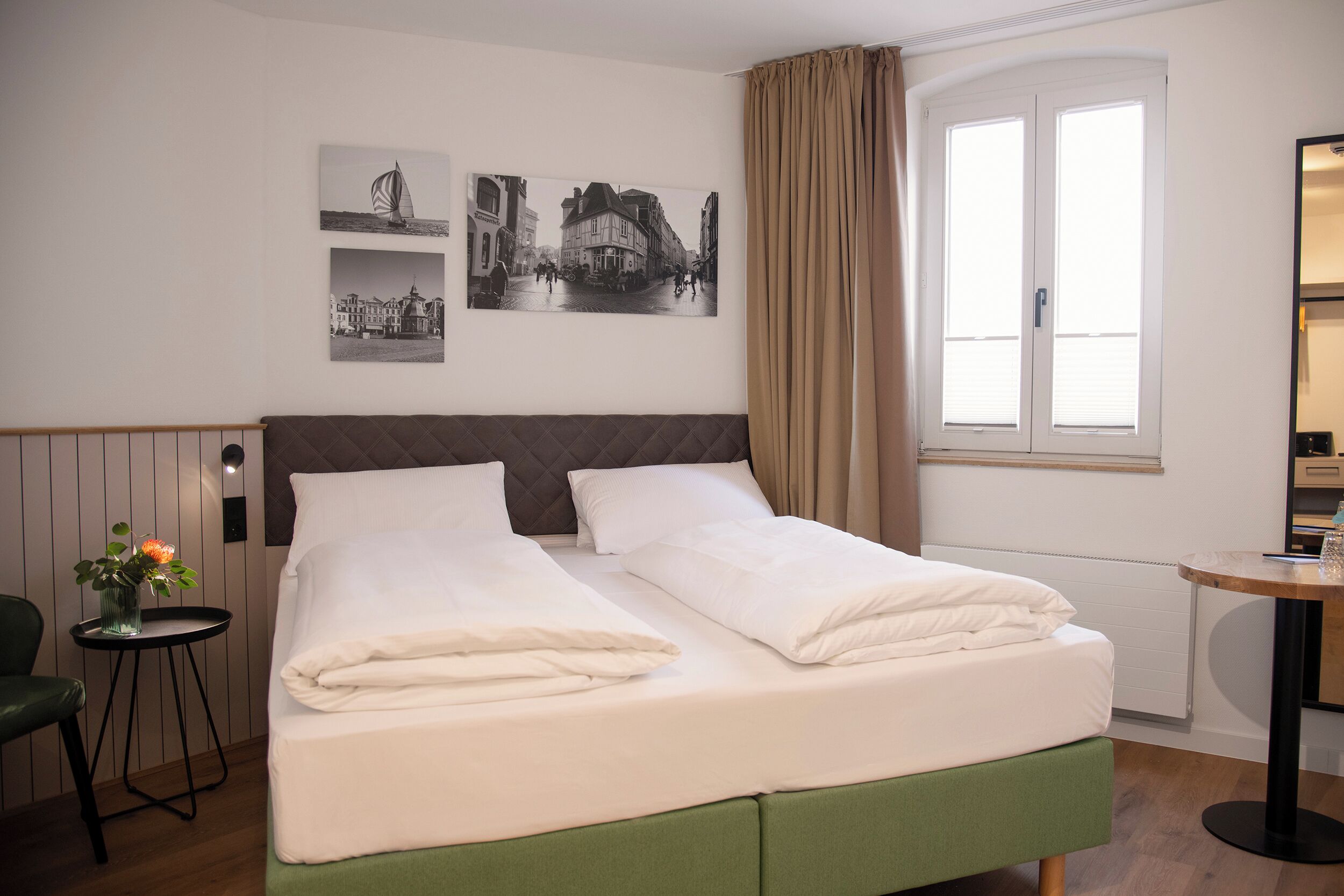 A neatly made bed with white linens, a green upholstered headboard, and a small table with a vase of flowers.