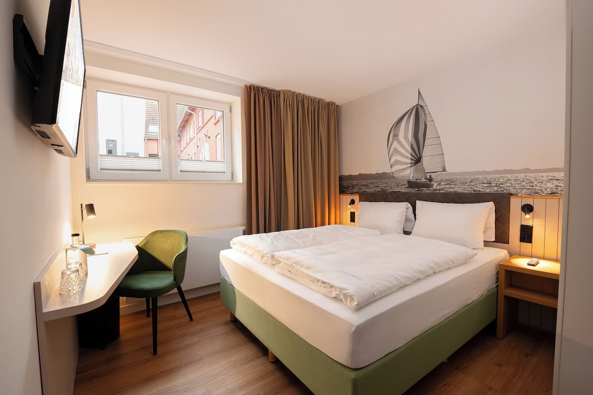 A hotel room with a bed, a desk, a chair, a window with curtains, a wall-mounted TV, and a wall art of a sailboat.