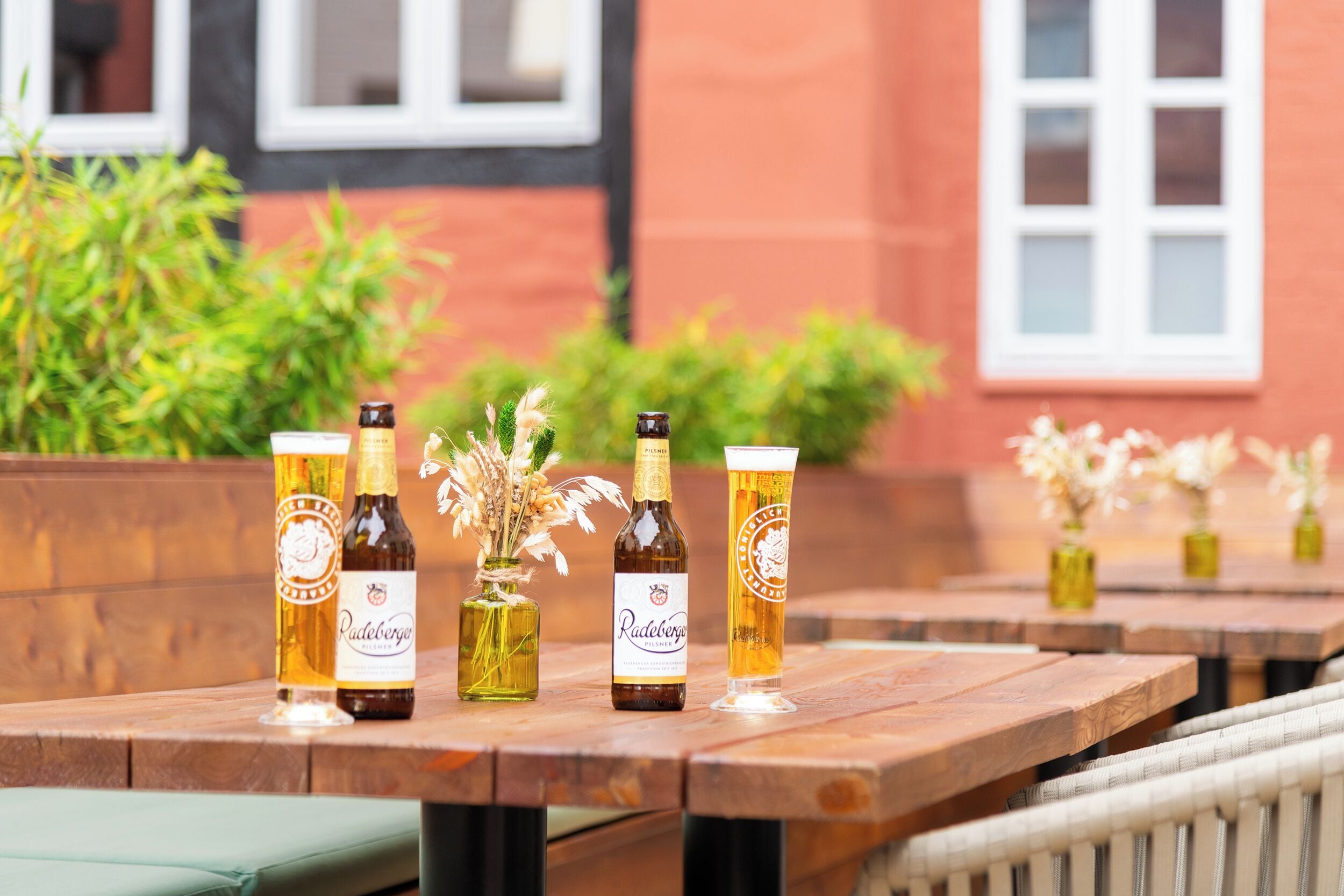 A row of wooden tables with beer bottles and glasses, a vase with dried flowers, and a building with windows in the background.