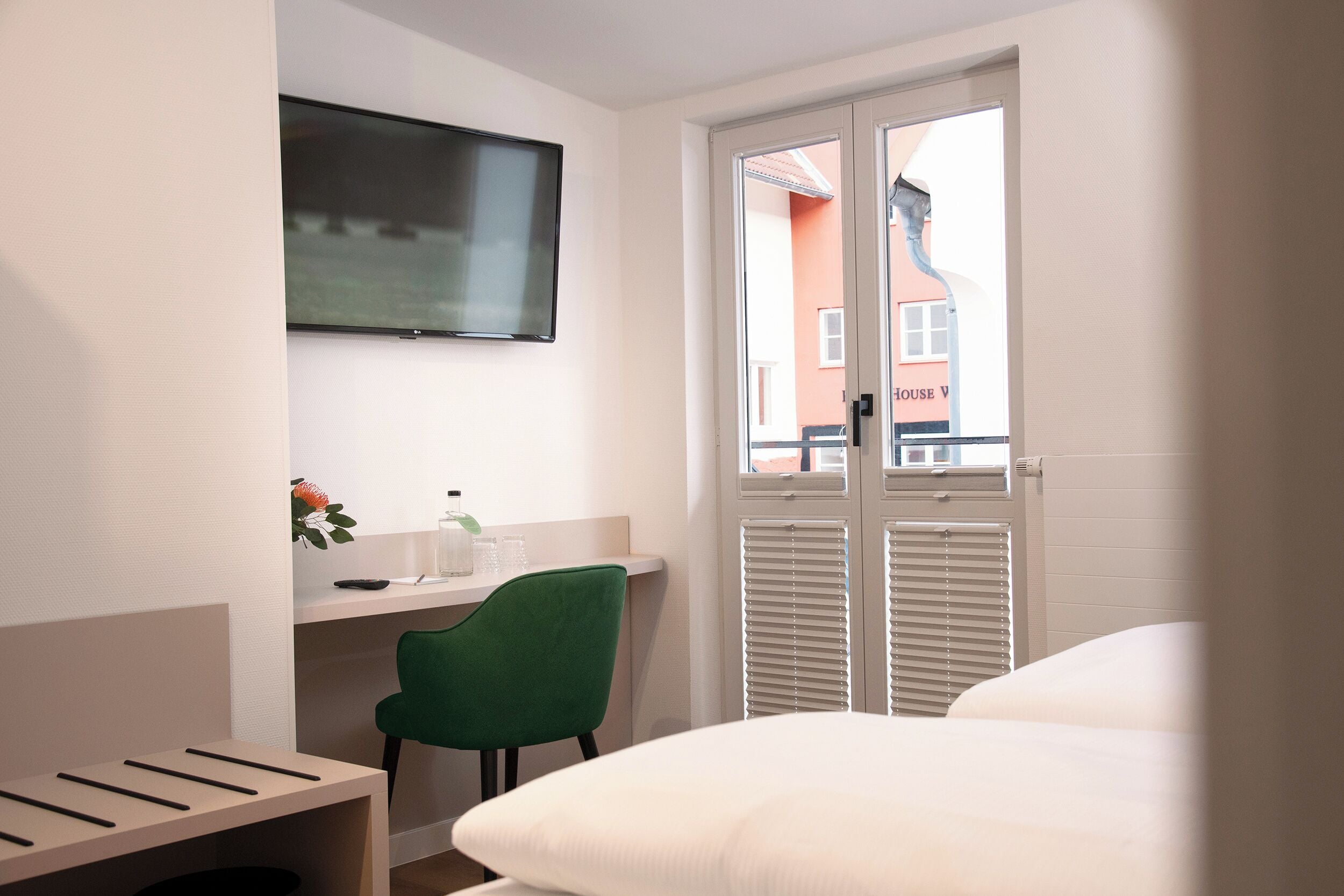Una habitación de hotel con cama, escritorio con silla, televisor montado en la pared y ventana con persianas.