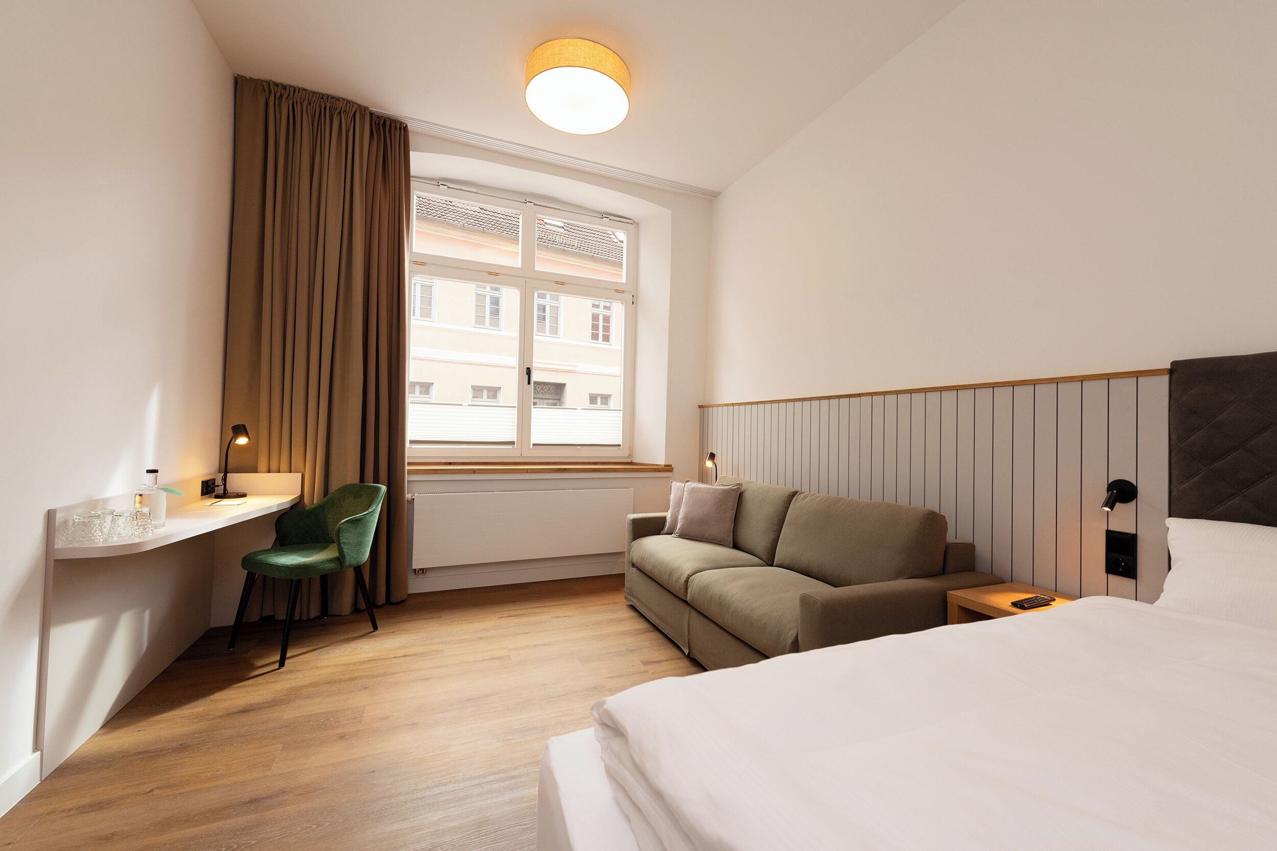 A hotel room with a bed, a desk with a chair, a sofa, and a window with curtains.