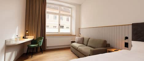 A hotel room with a bed, a desk with a chair, a sofa, and a window with curtains.