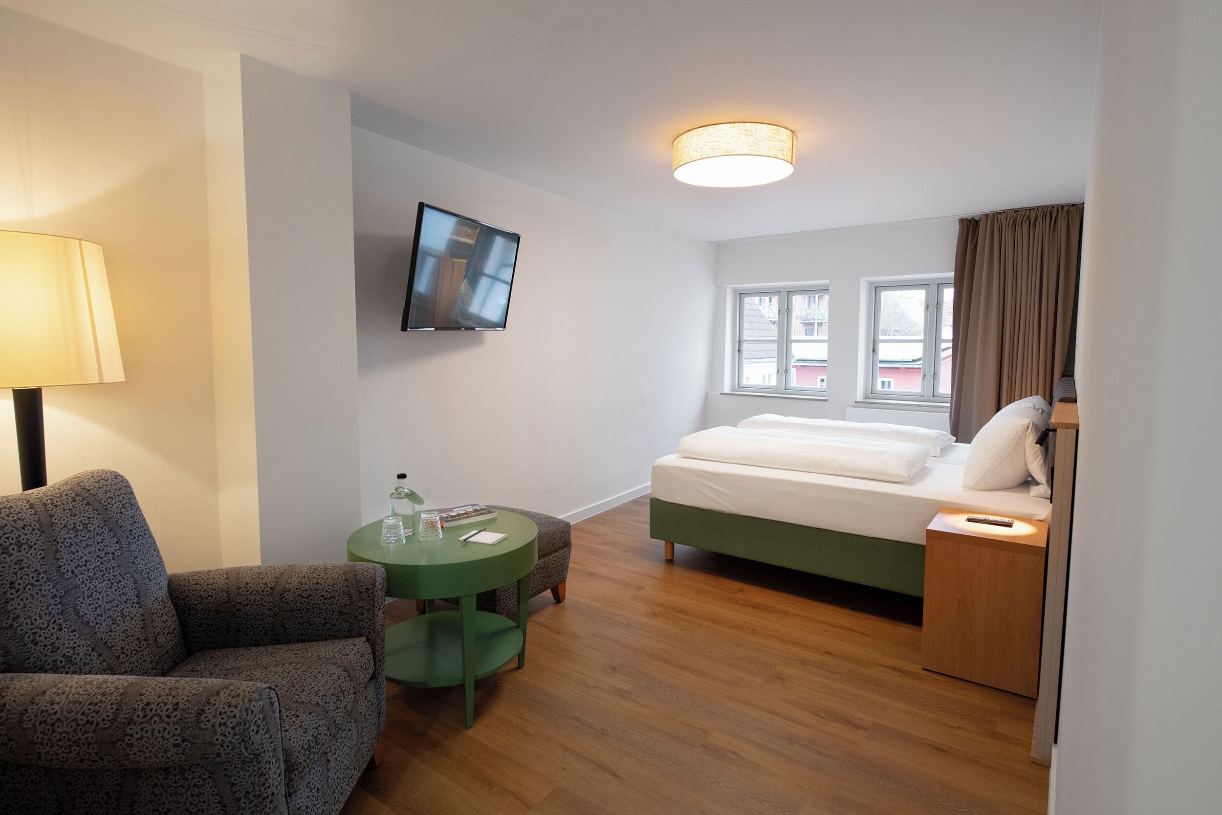 A hotel room with a bed, a TV mounted on the wall, a green side table, a lamp, and a window with curtains.
