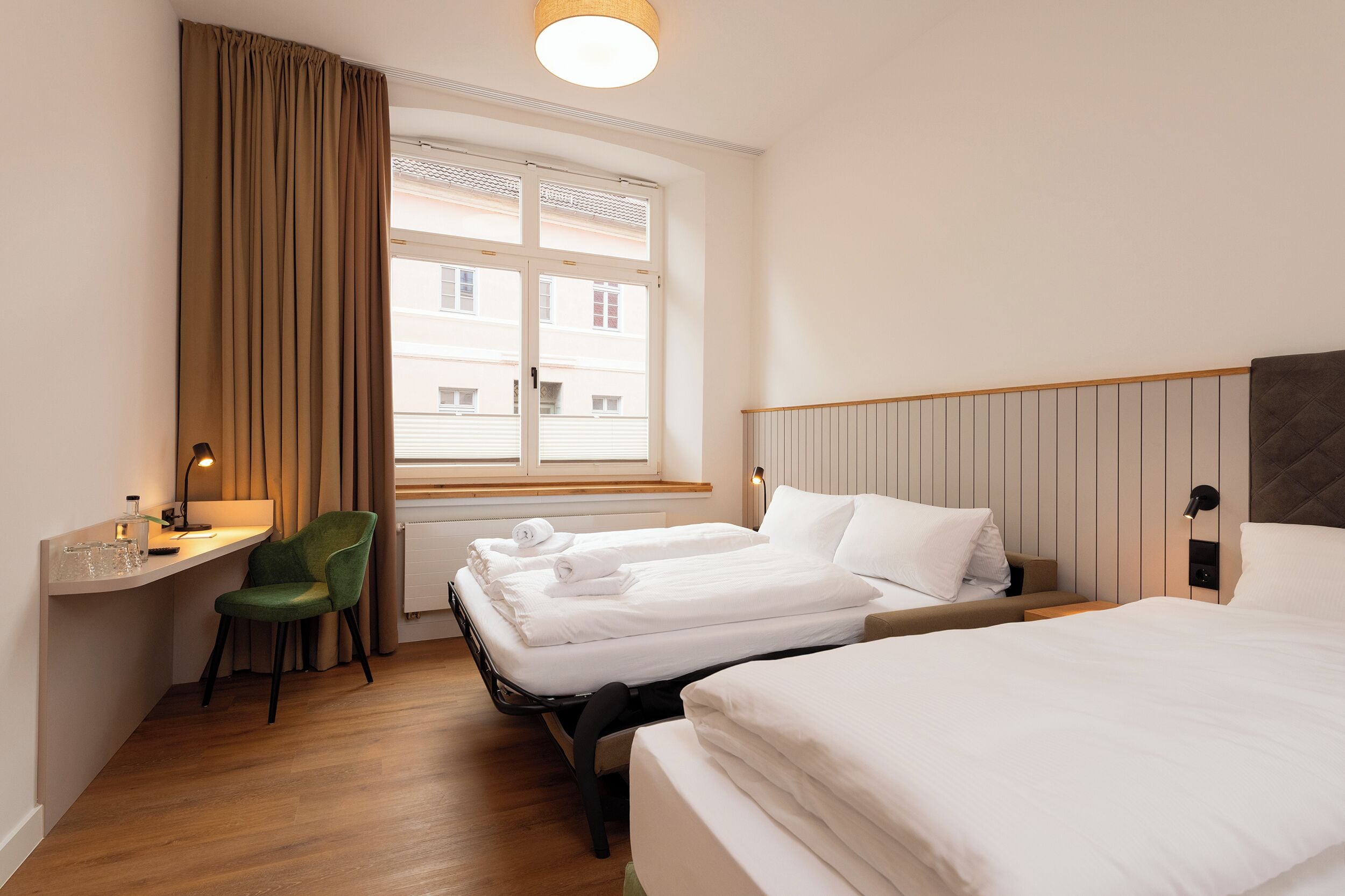 Habitación de hotel con dos camas, un escritorio, una silla, un ventanal con cortinas y una mesita de noche con lámpara.