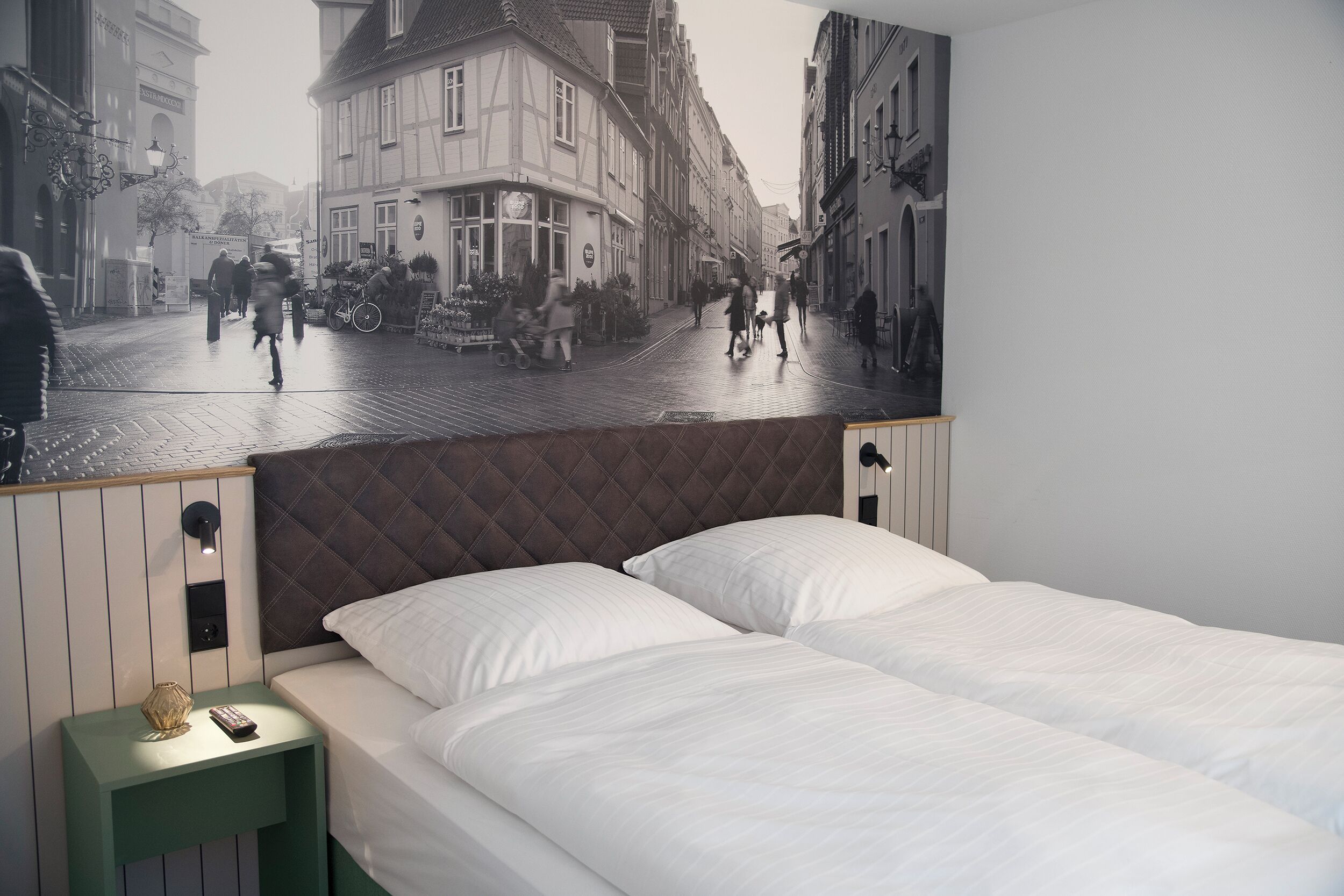 Habitación de hotel con cama, mesita de noche y un mural en la pared que representa una escena callejera.