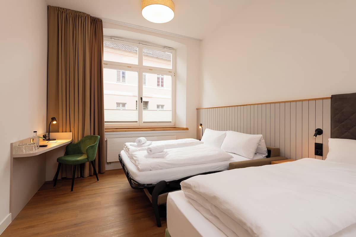 A hotel room with two beds, a desk, a chair, a window with curtains, and a bedside table with a lamp.