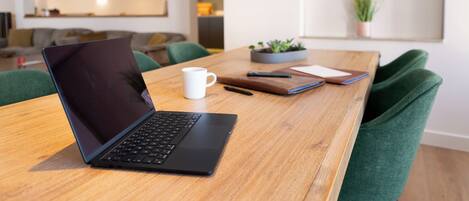 A laptop on a wooden table with a white mug, a notebook, and a pen. There is a green couch and a plant in the background.