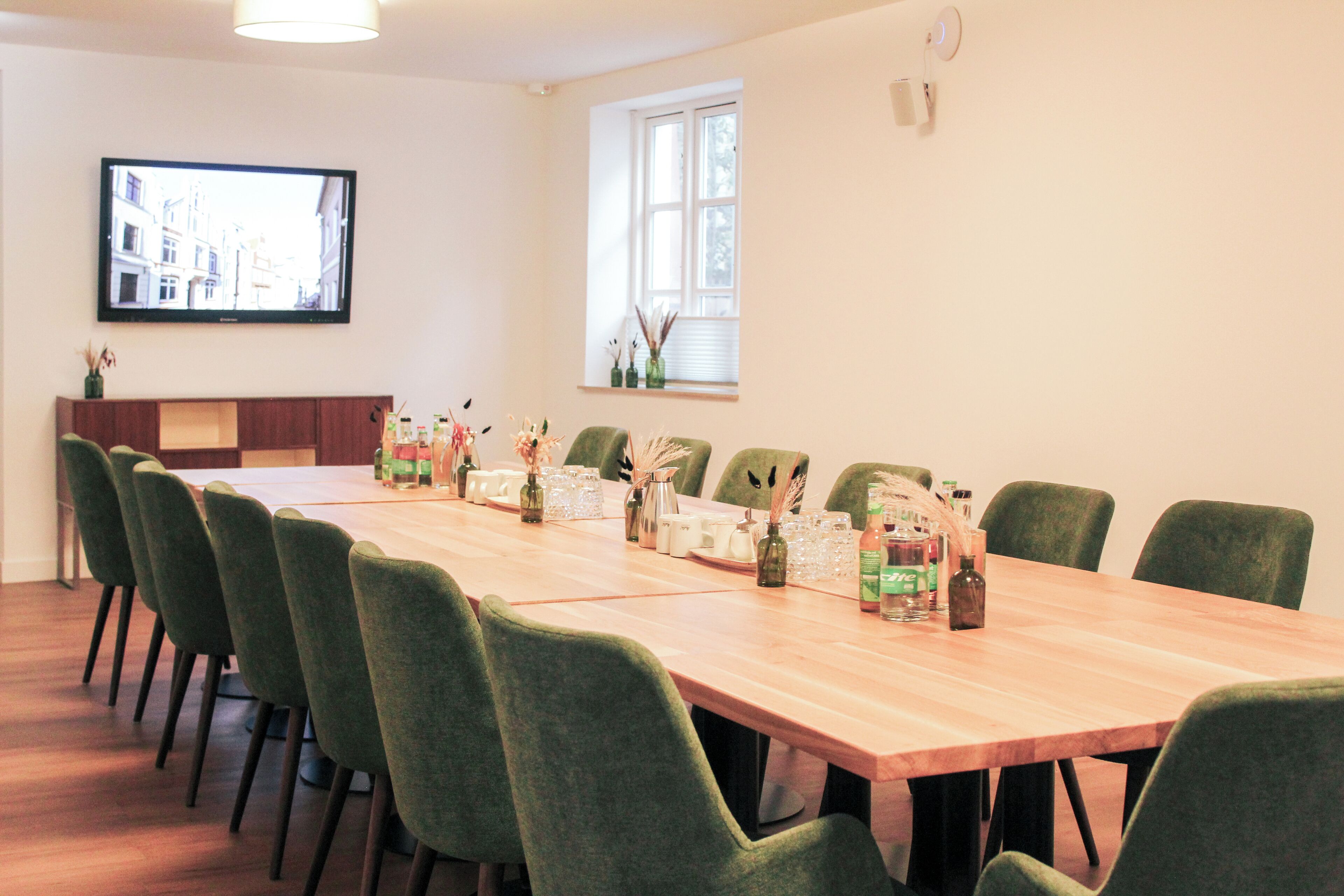 A conference room with a long table, green chairs, a television on the wall, and a window with blinds.