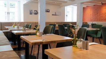 Een restaurant met houten tafels en groene stoelen, een bakstenen muur en kunst aan de wanden.