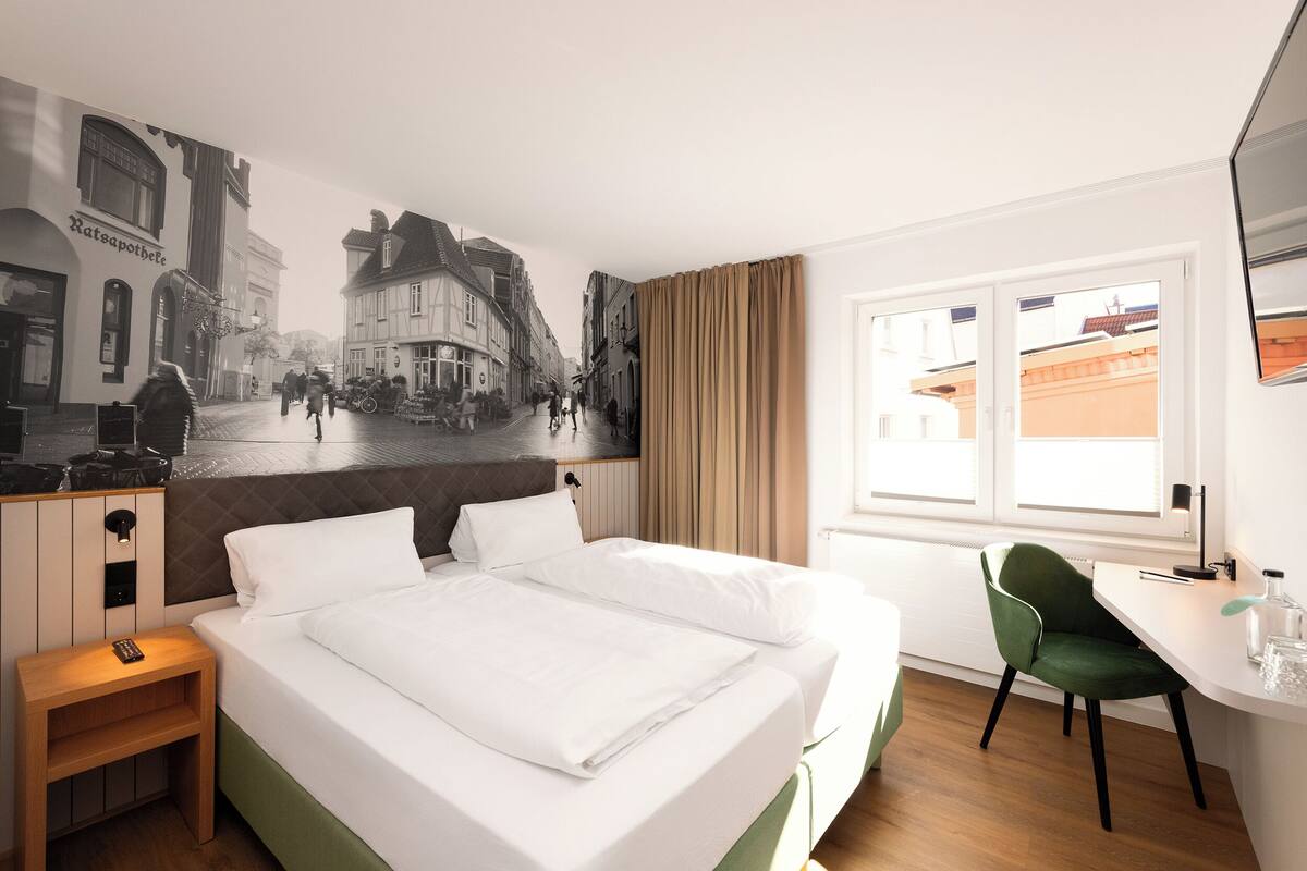 A hotel room with a bed, a desk, a chair, a wall mural, and a window.