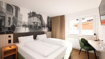 Una habitación de hotel con cama, escritorio, silla, un mural en la pared y ventana.