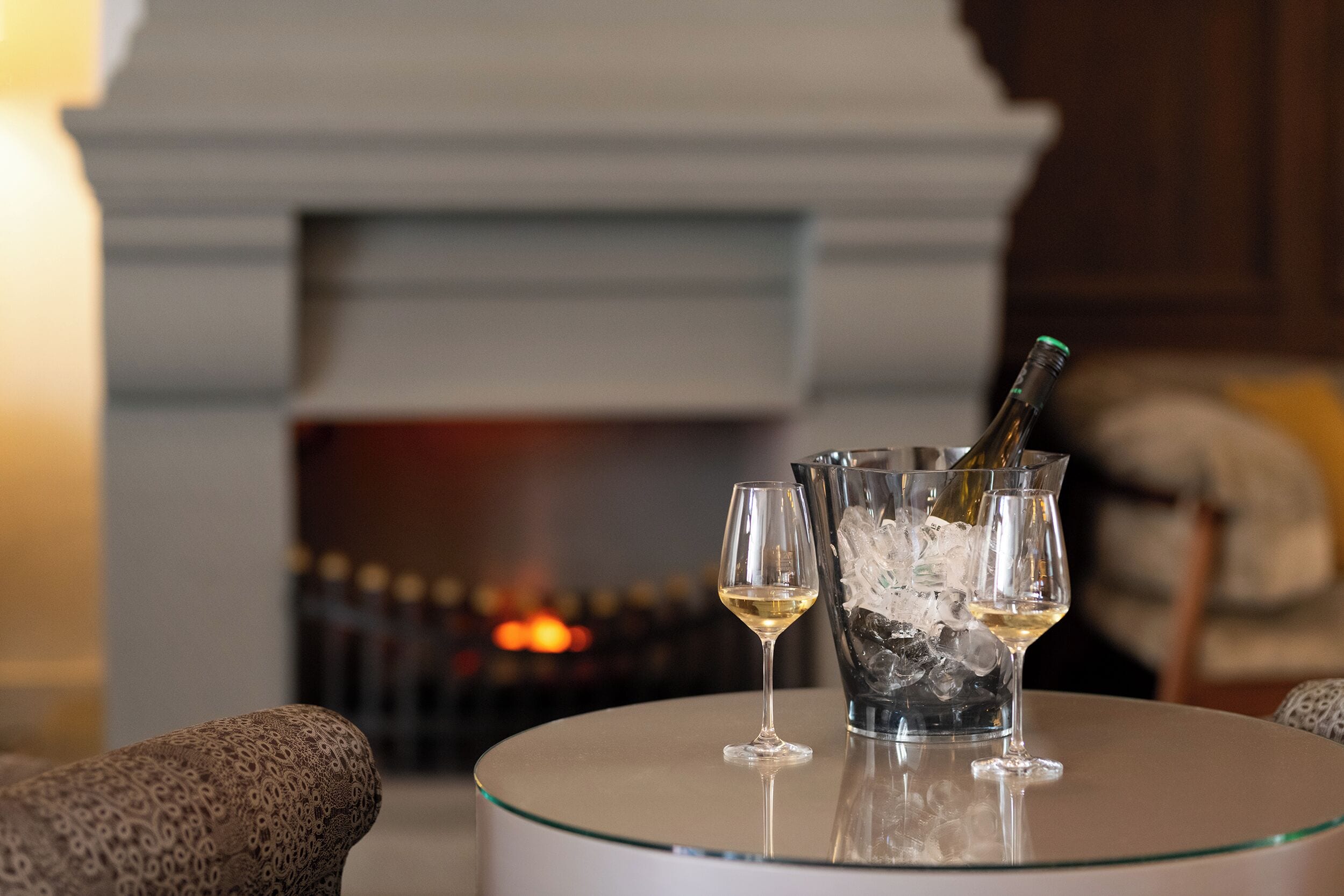 A fireplace with a fire, a table with a bottle of champagne and two glasses, and patterned armchairs.