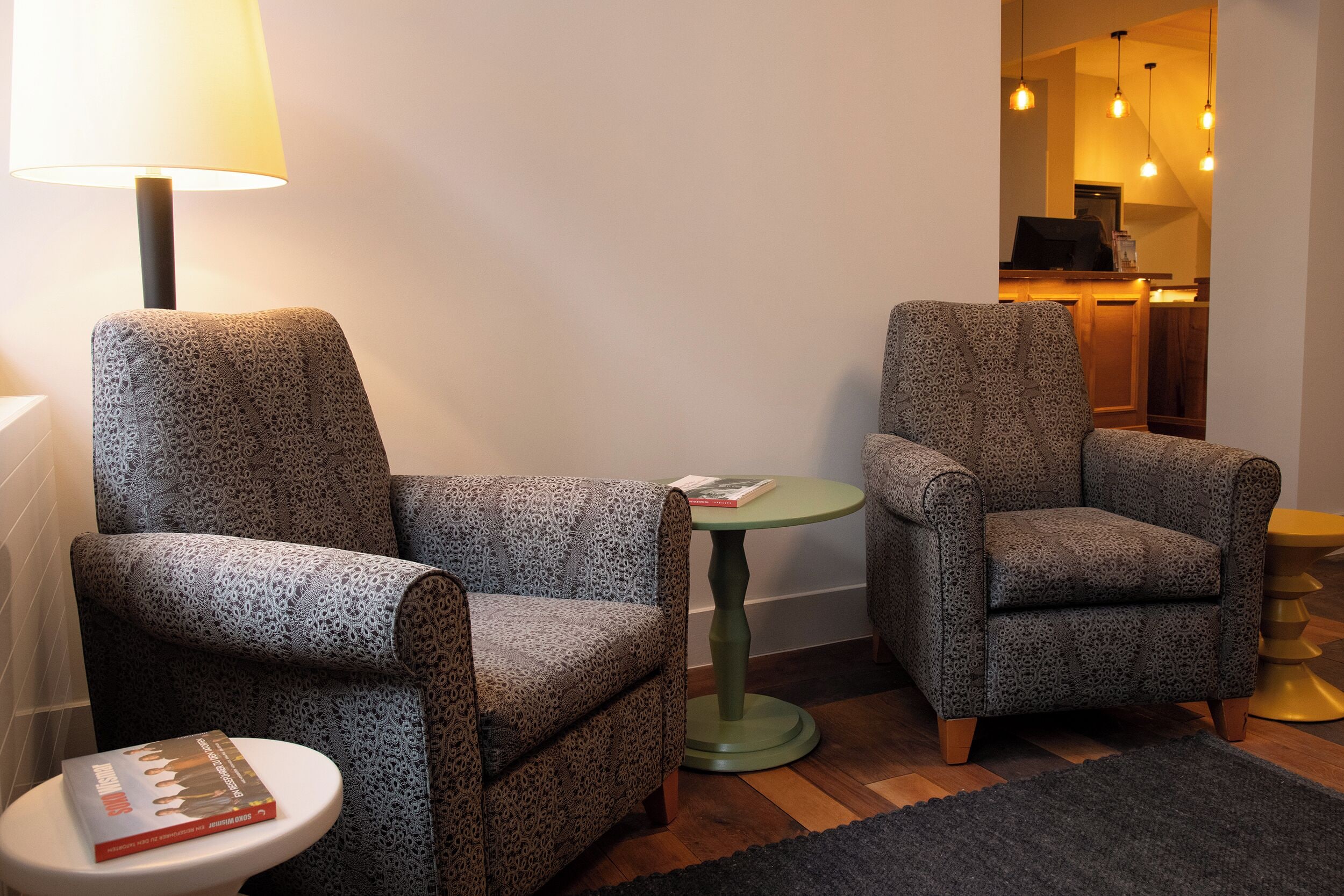 A small, cozy room with two patterned armchairs, a small round table, a lamp, and a book on a side table.