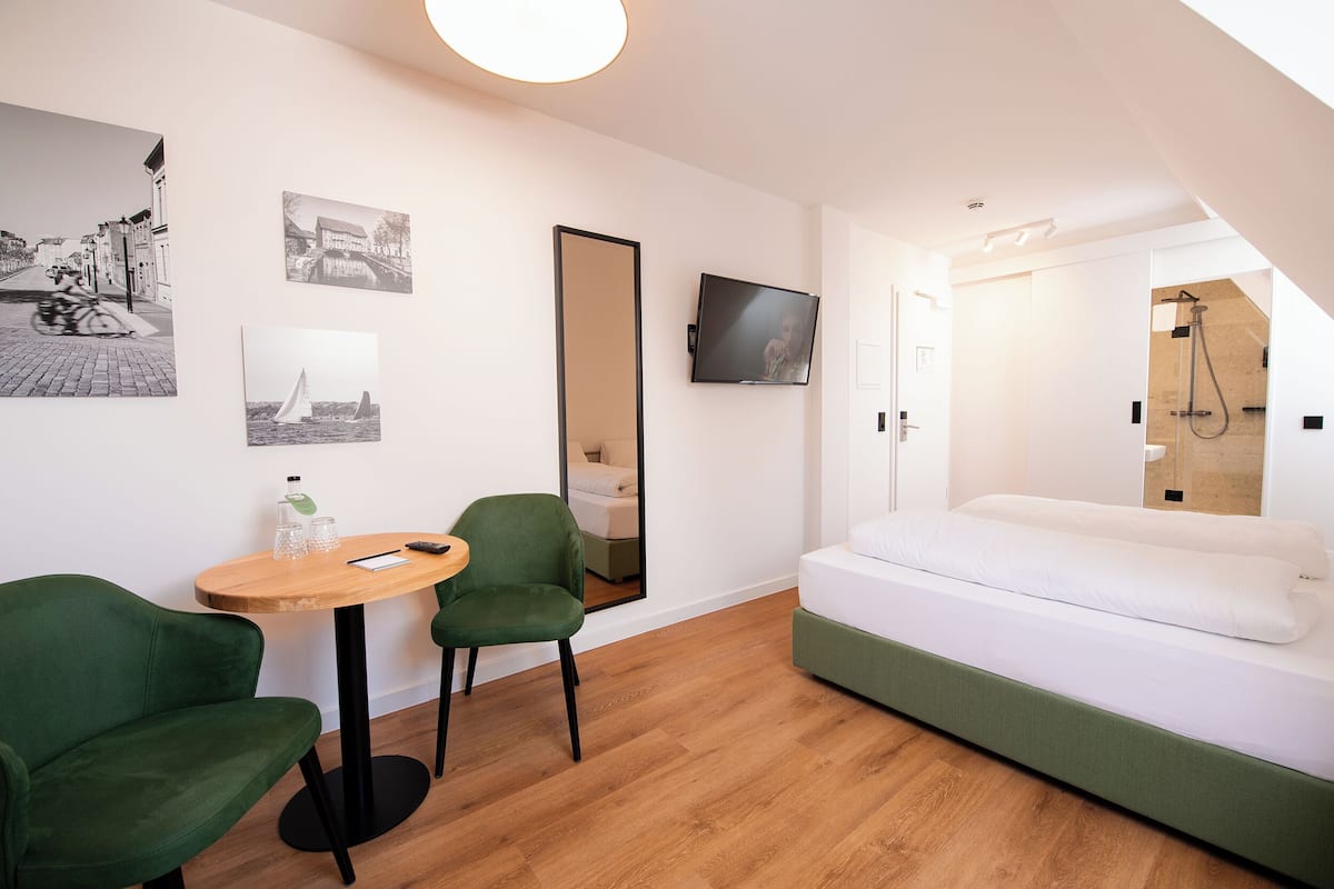 A modern hotel room with a wooden floor, a green sofa, a small round table with chairs, a bed with white bedding, and a TV mounted on the wall.