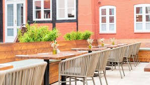 Un espace extérieur aménagé avec des tables et des chaises en bois, un bac à fleurs orné de plantes vertes et, en arrière-plan, un bâtiment dont les fenêtres sont encadrées de blanc.