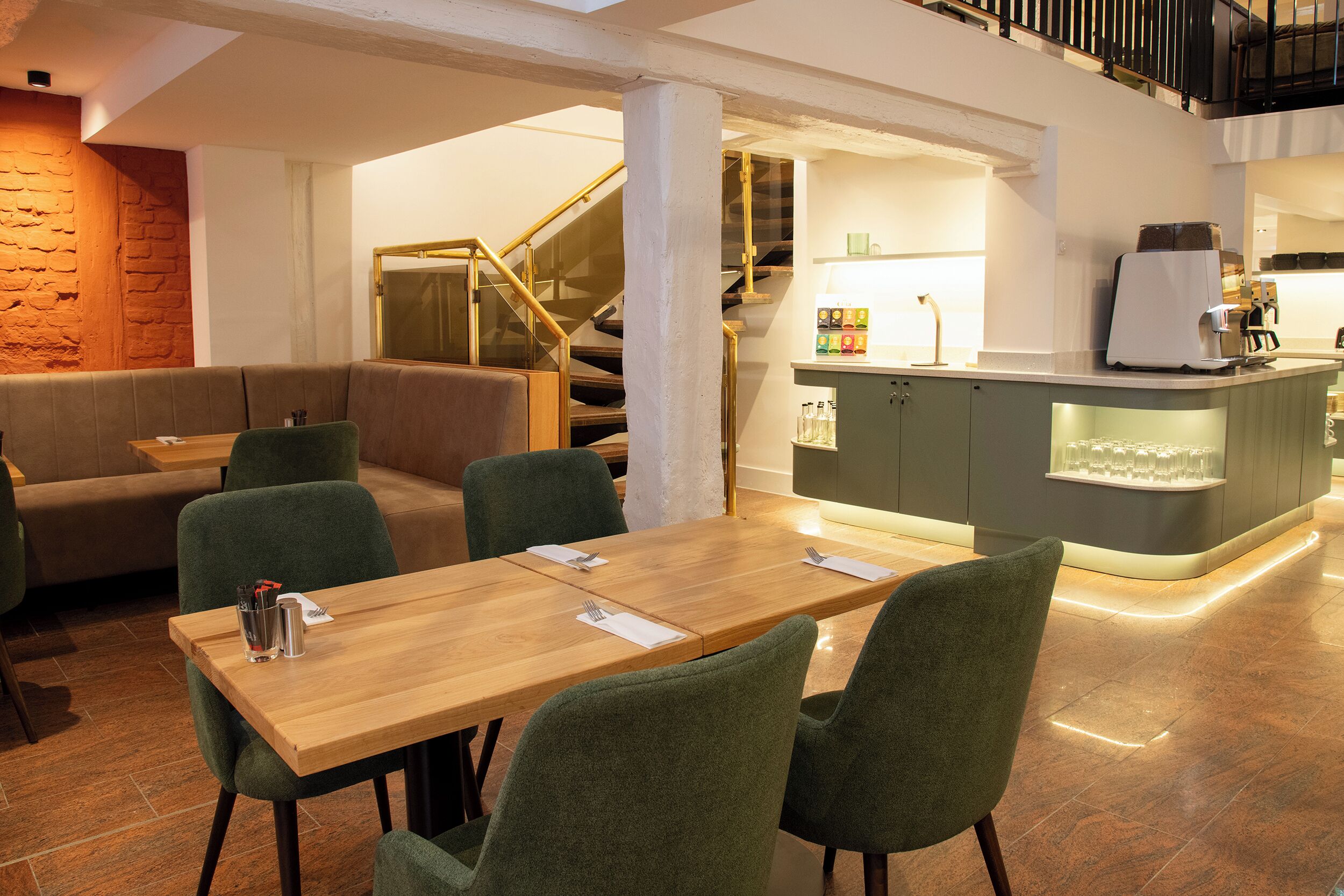 Un comedor moderno con una mesa de madera y sillas verdes, una zona de bar con mostrador y taburetes, y una escalera que lleva a un nivel superior.