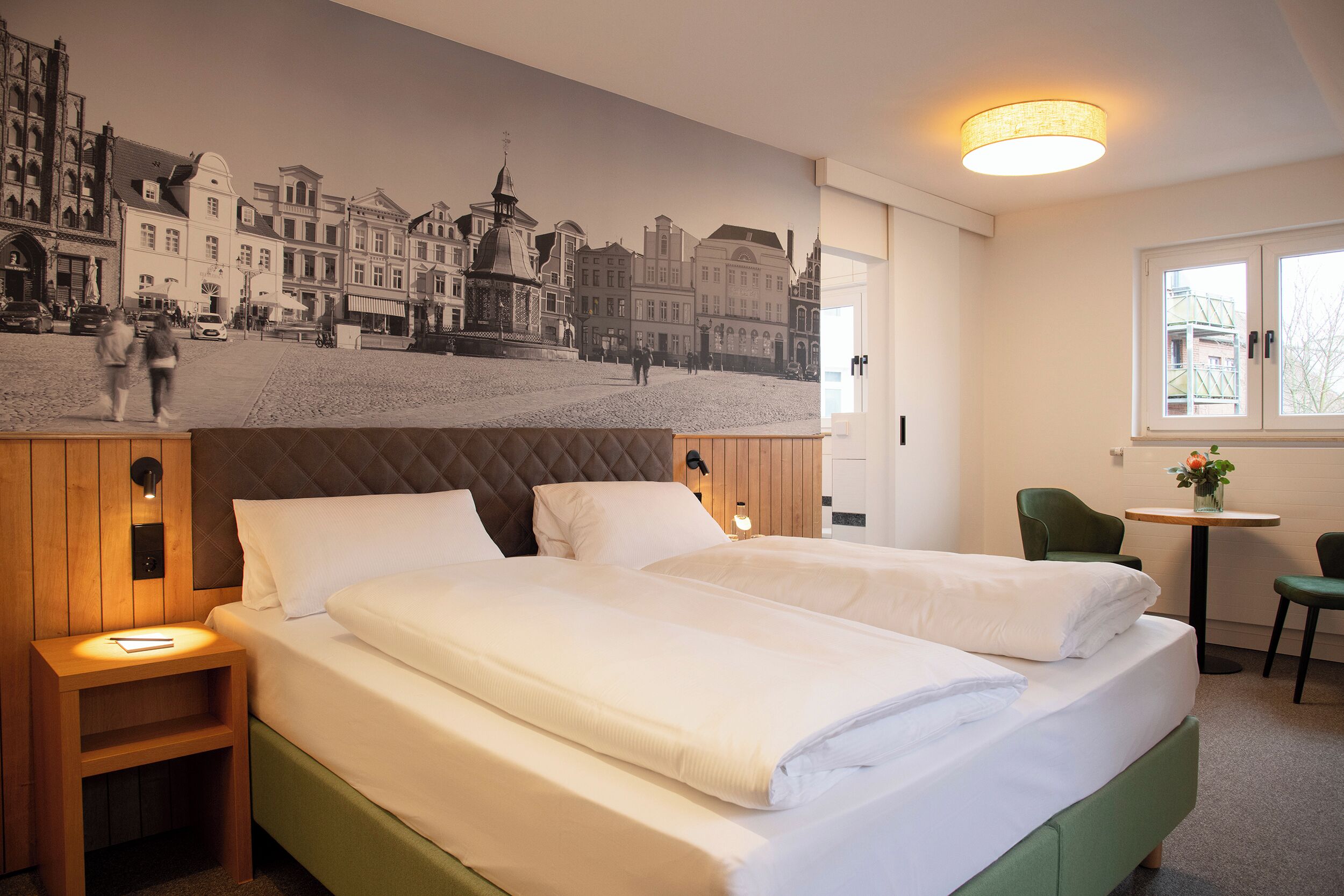 A hotel room with two beds, a mural of a cityscape, a small table with a chair, and a window with a view.