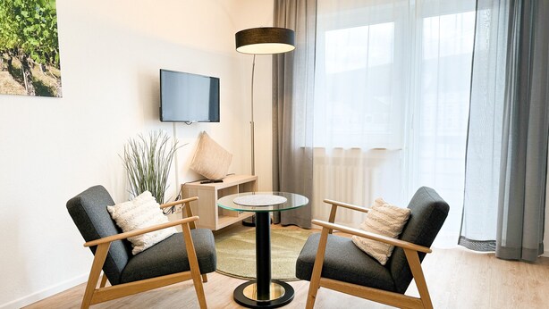 A modern living room with two armchairs, a round coffee table, a TV mounted on the wall, and a large window with sheer curtains.