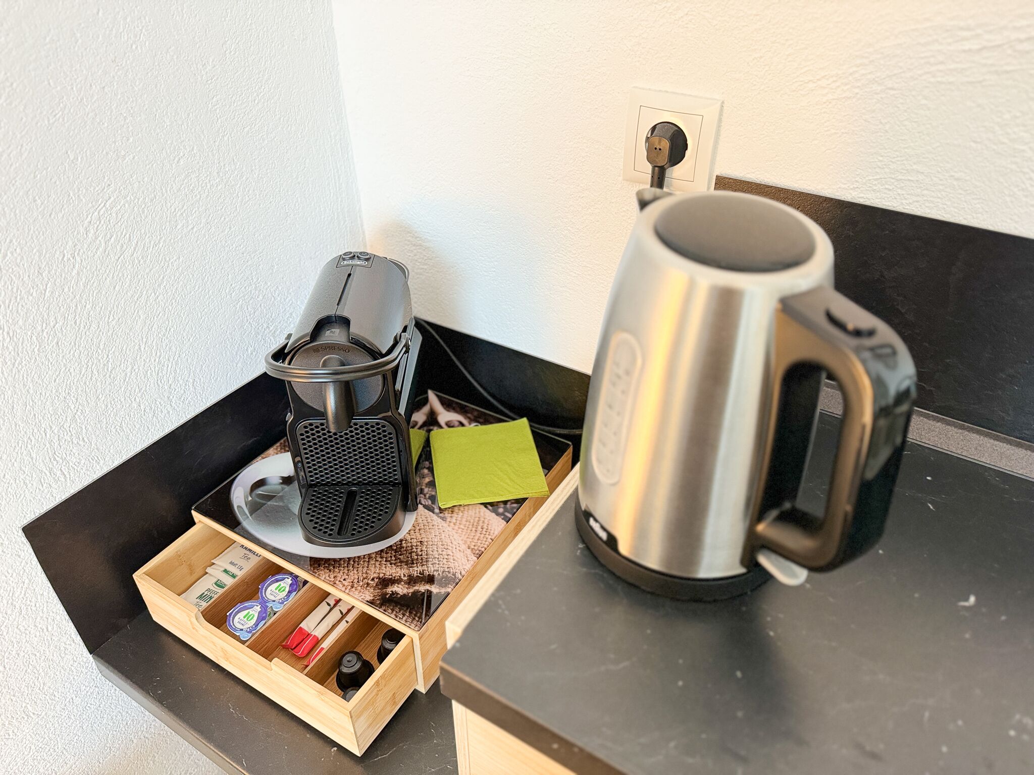 A coffee maker, a kettle, and a wooden drawer with coffee packets on a countertop.