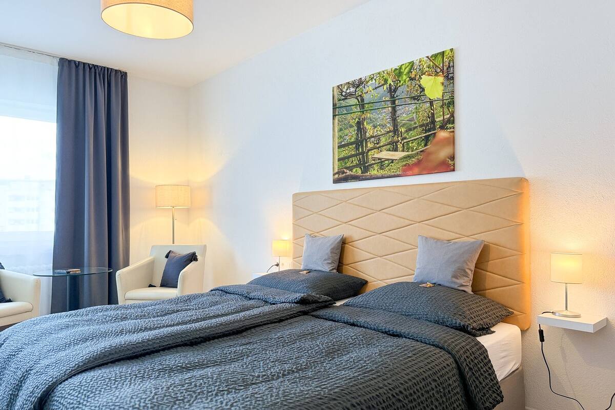 A modern hotel room with a large bed, two bedside tables with lamps, a chair, and a painting on the wall.