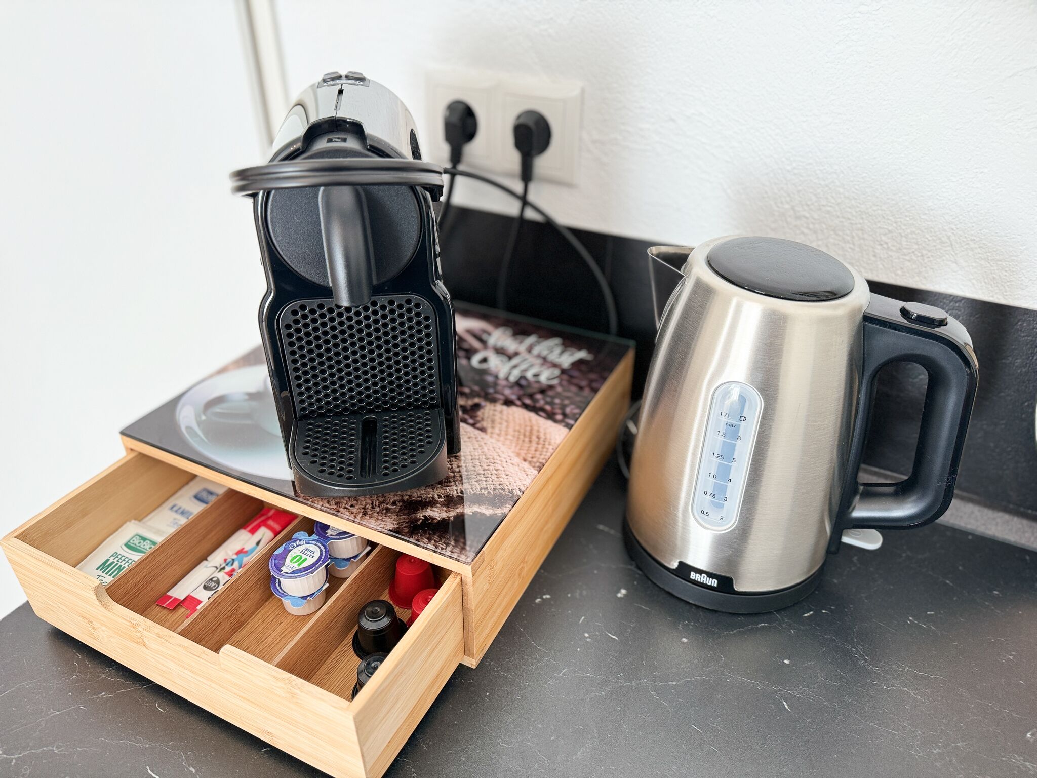 A coffee maker, a kettle, and a drawer with coffee pods and a red container on a kitchen counter.