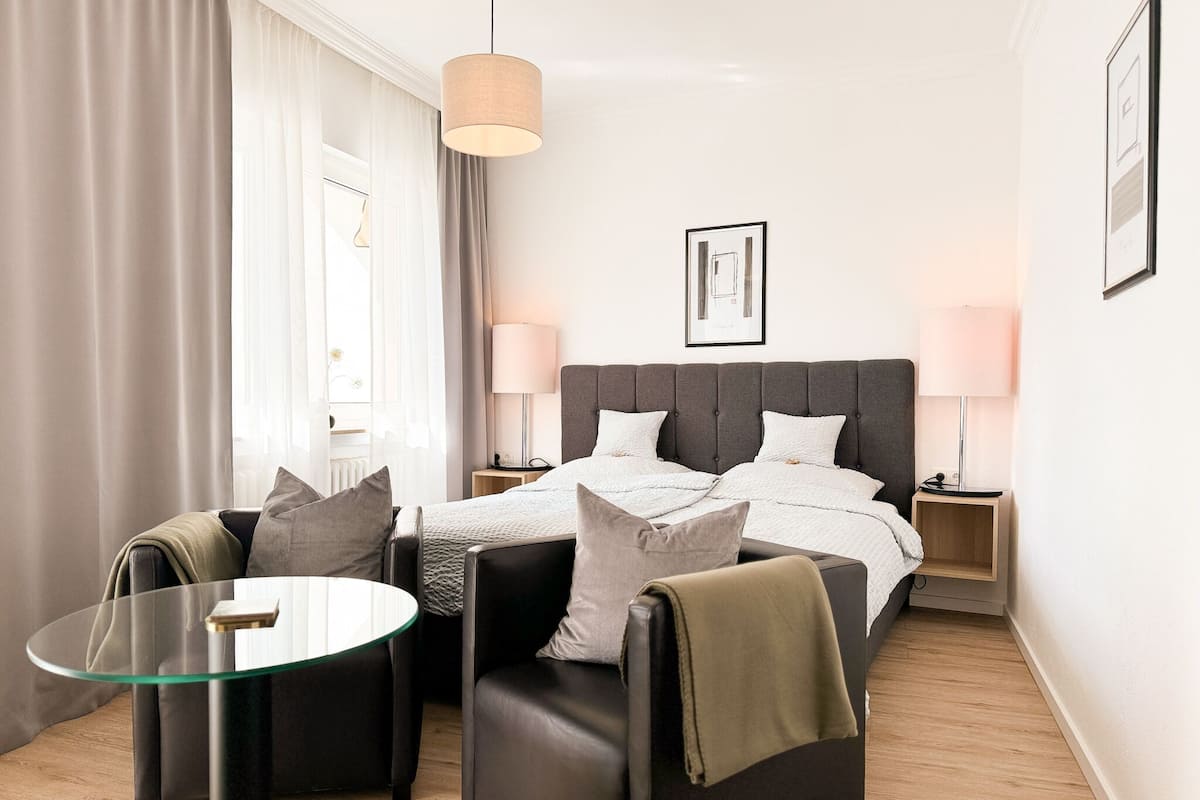 A modern hotel room with a bed, two bedside tables, a glass coffee table, and a seating area with two chairs.