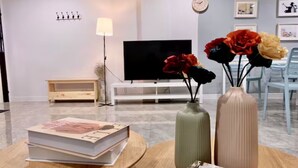 Ein modernes Wohnzimmer mit einem Flachbildfernseher, einem kleinen Holznachttisch und einem runden Couchtisch mit Büchern und Vasen.