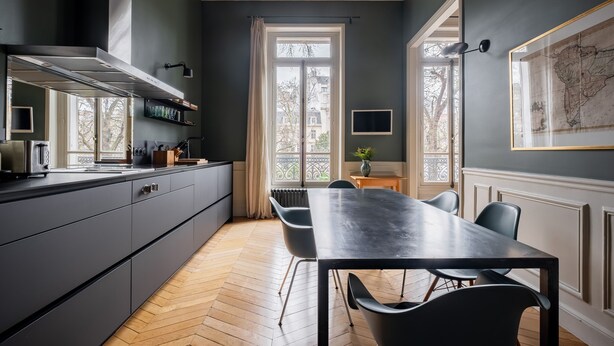 A modern kitchen with dark cabinetry, a large dining table, and a view through a window to the outside. - Augustin Glory, by Veeve (Paris)