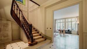 A grand staircase with a wooden handrail and beige steps, leading to a room with a large window and a dining area. - Augustin Glory, by Veeve (Paris)