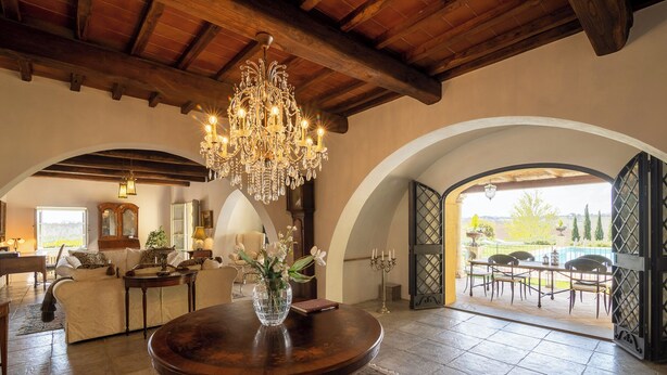 Ein geräumiges Wohnzimmer mit einer Chandelüstere, einem runden Holztisch und Blick durch einen Bogen zur Terrasse.
