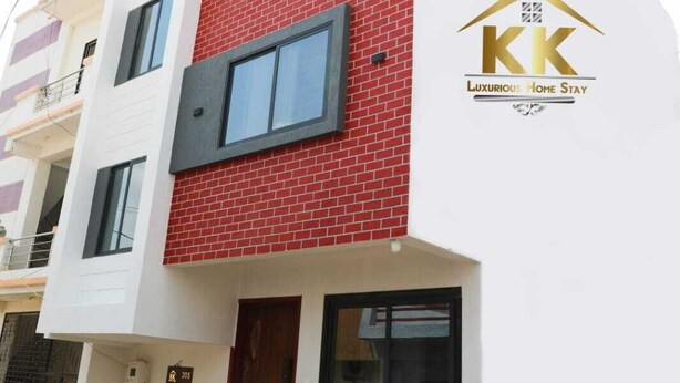 A multi-story building with a red brick facade and white walls. There is a logo with the letters "KK" and the words "Luxury Home Stay" on the right side of the building. - K K Luxurious Home Stay (Garudeshwar)