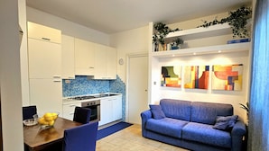 A modern living room with a blue sofa, a dining table with chairs, and a kitchen area with white cabinets and a tiled backsplash. - Beautiful two-bedroom apartment in a central location, just a few steps from the sea   (Giardini Naxos)
