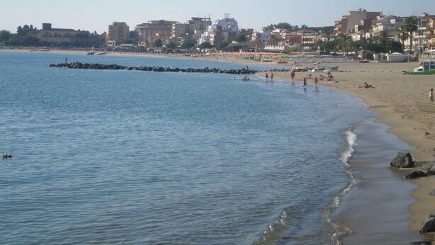 A beach with a rocky breakwater, clear blue water, and a cityscape in the background. - Beautiful two-bedroom apartment in a central location, just a few steps from the sea   (Giardini Naxos)