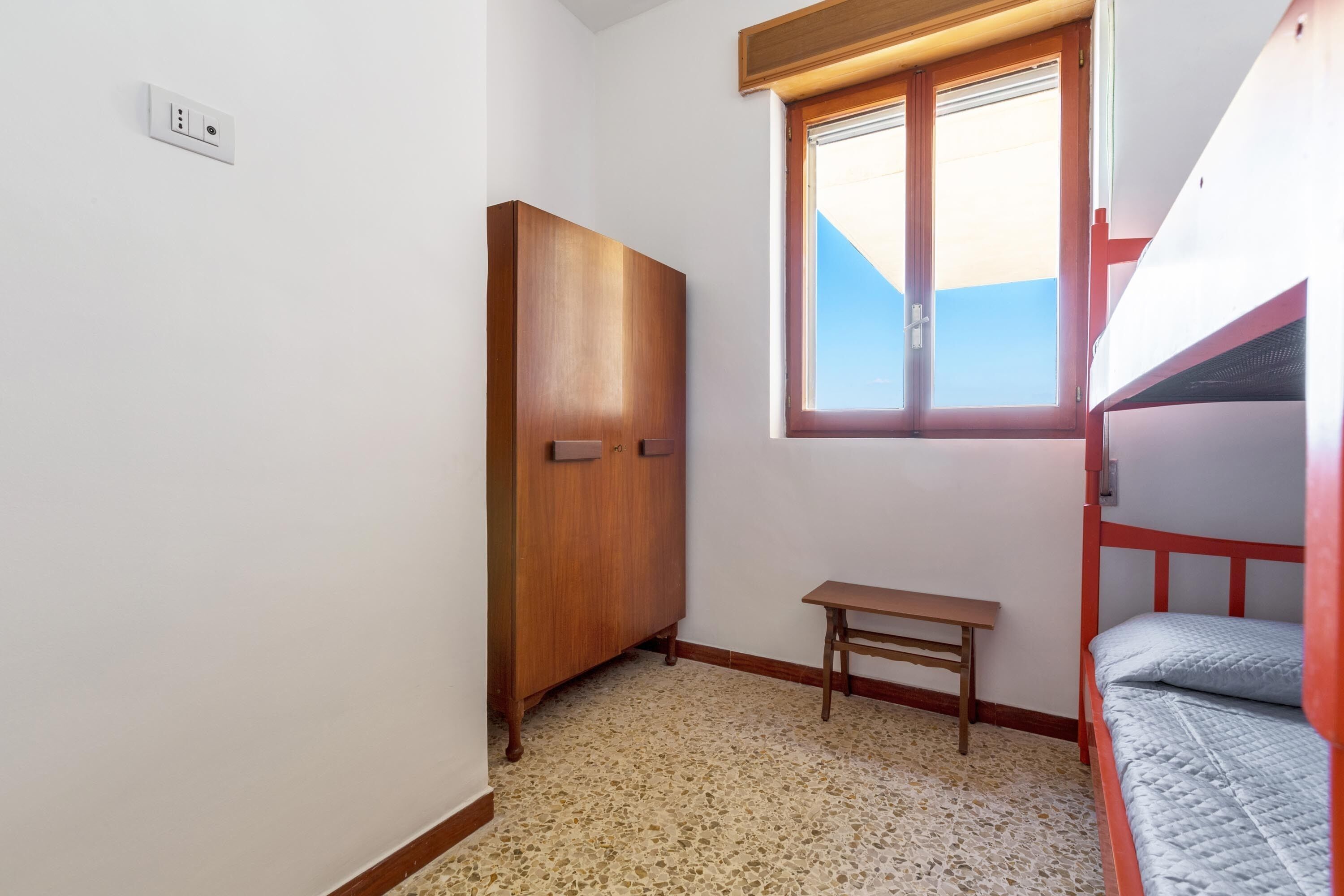 A room with a wooden wardrobe, a small wooden table, a bunk bed, and a window with a view.