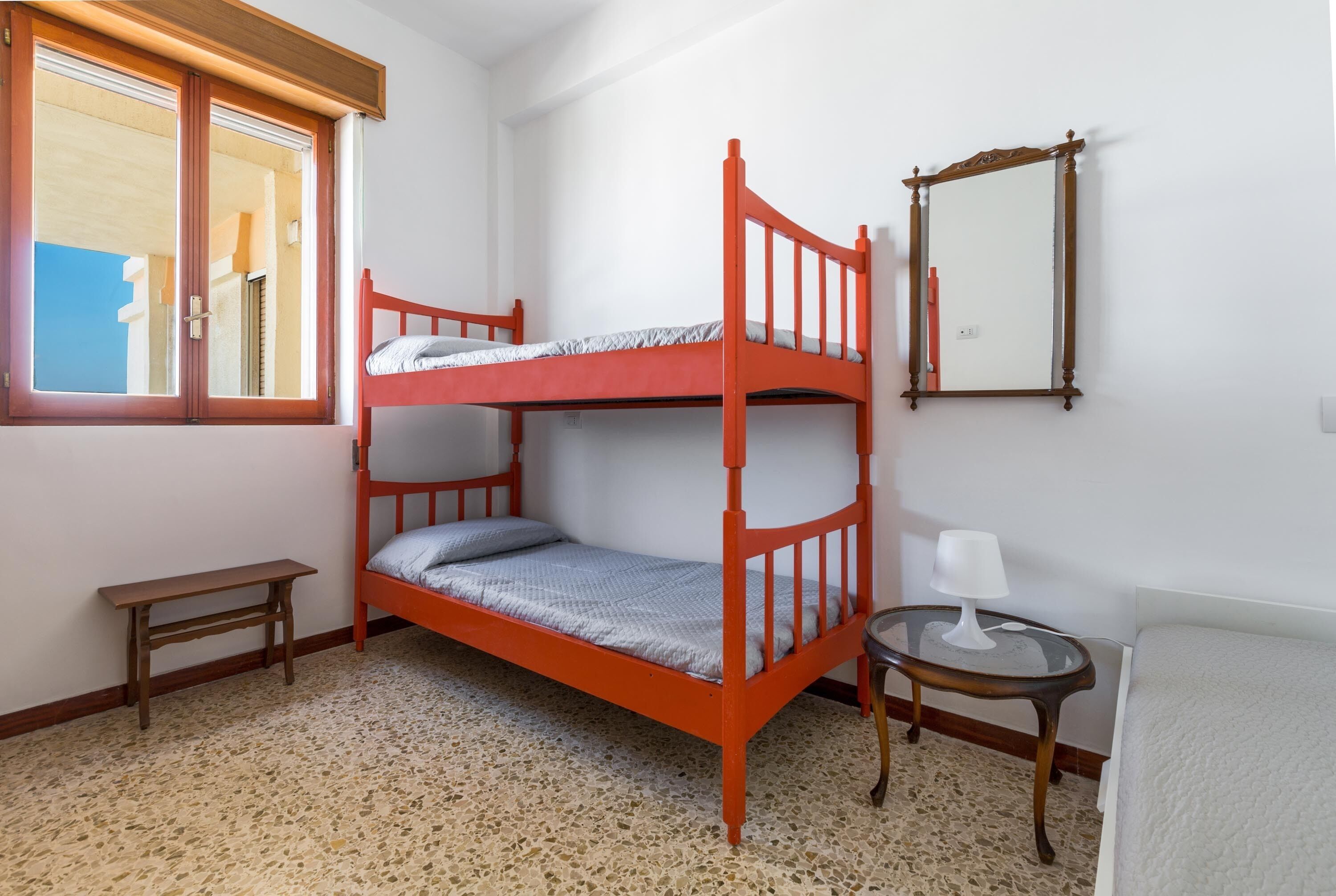 A bunk bed room with a window, a small table with a lamp, and a mirror on the wall.
