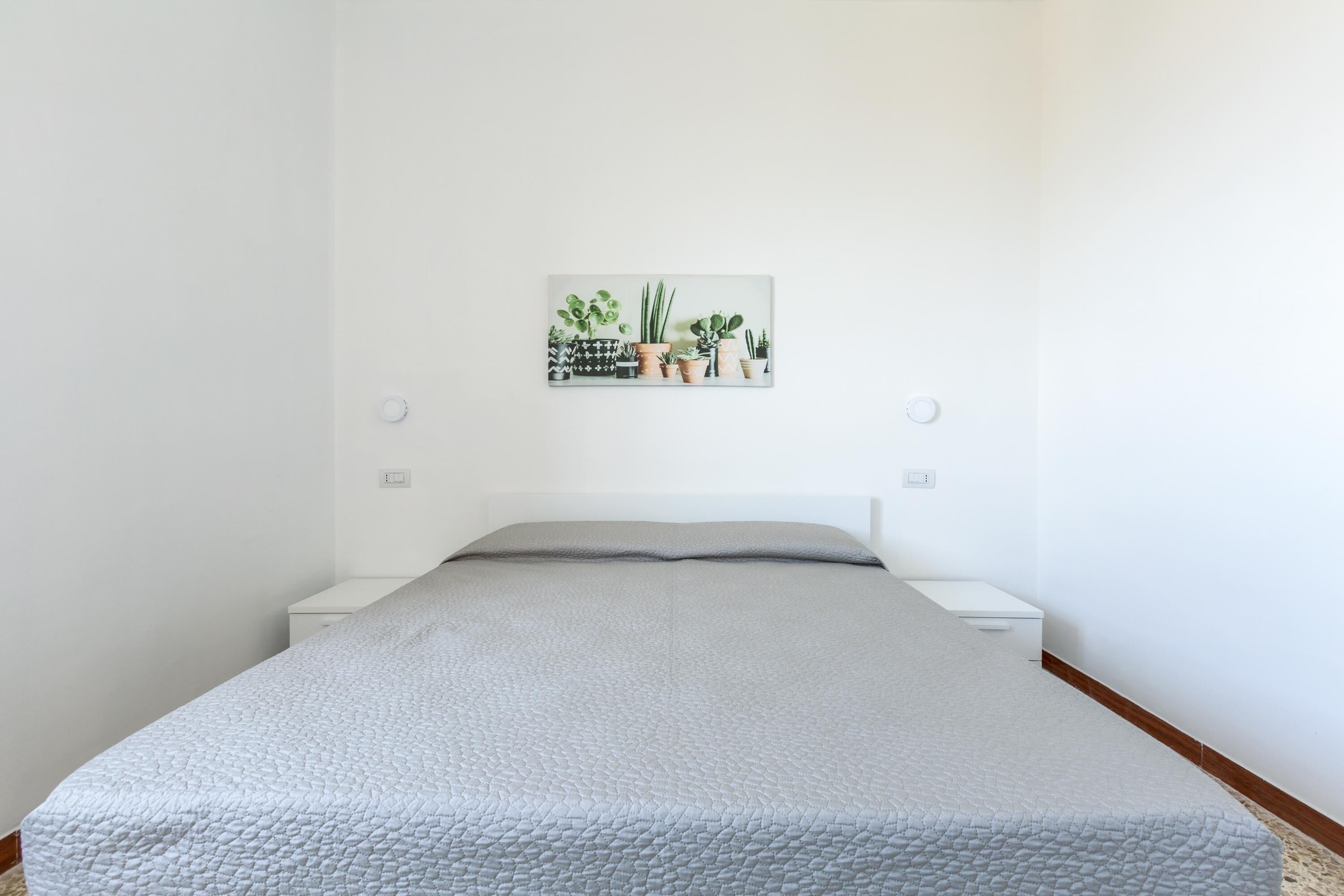 Een slaapkamer met een bed, witte nachtkastjes, een aan de muur bevestigde plant en een klein wit kastje.