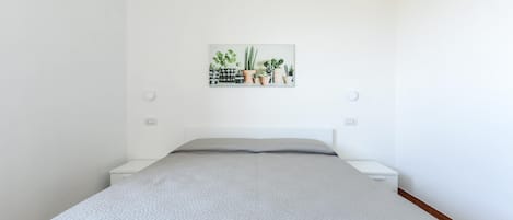 Een slaapkamer met een bed, witte nachtkastjes, een aan de muur bevestigde plant en een klein wit kastje.