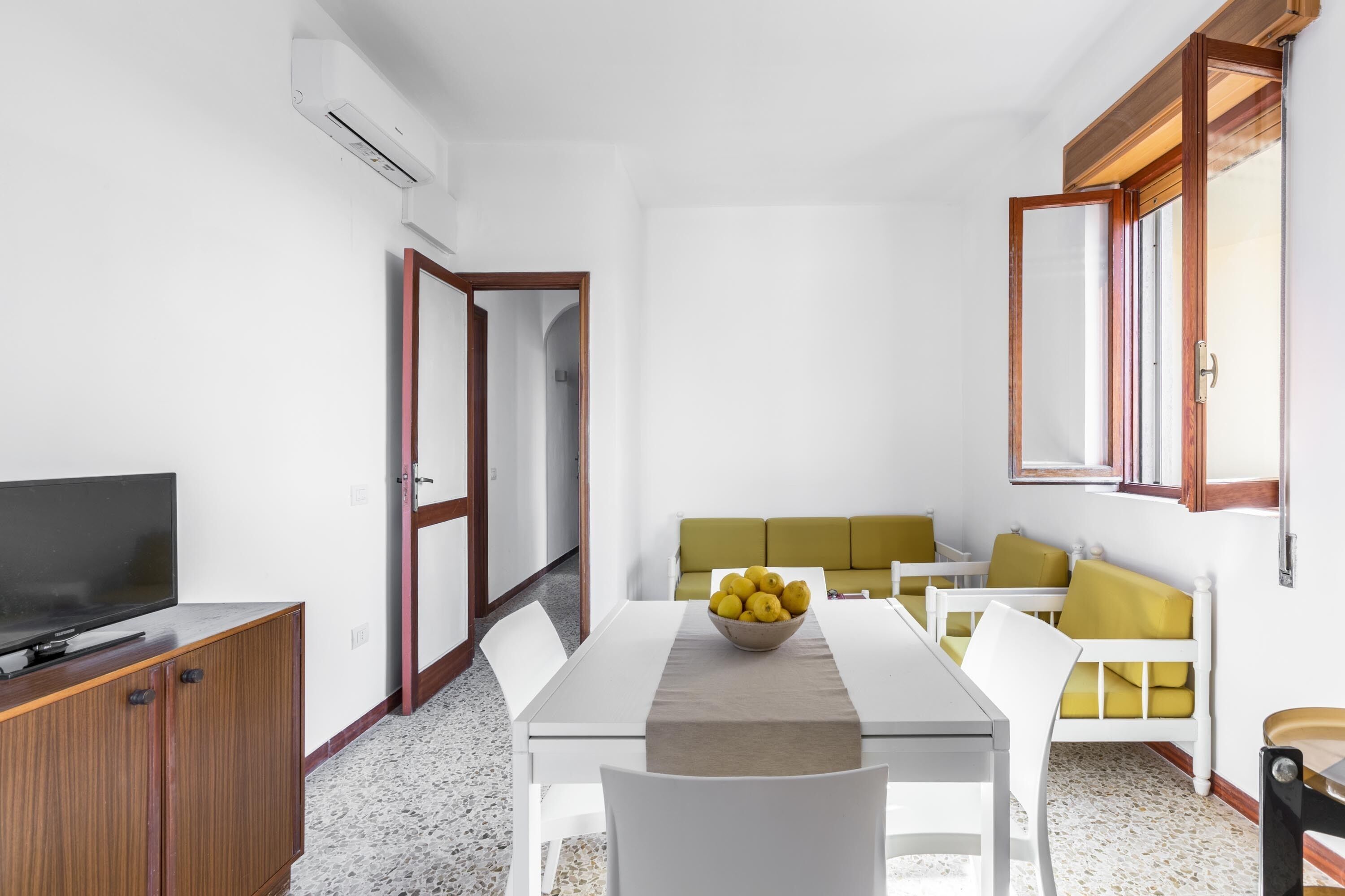 Ein modernes Wohnzimmer mit Fernseher, einem Esstisch mit Obstschale, einer Couch und einem Fenster mit Holzrahmen.