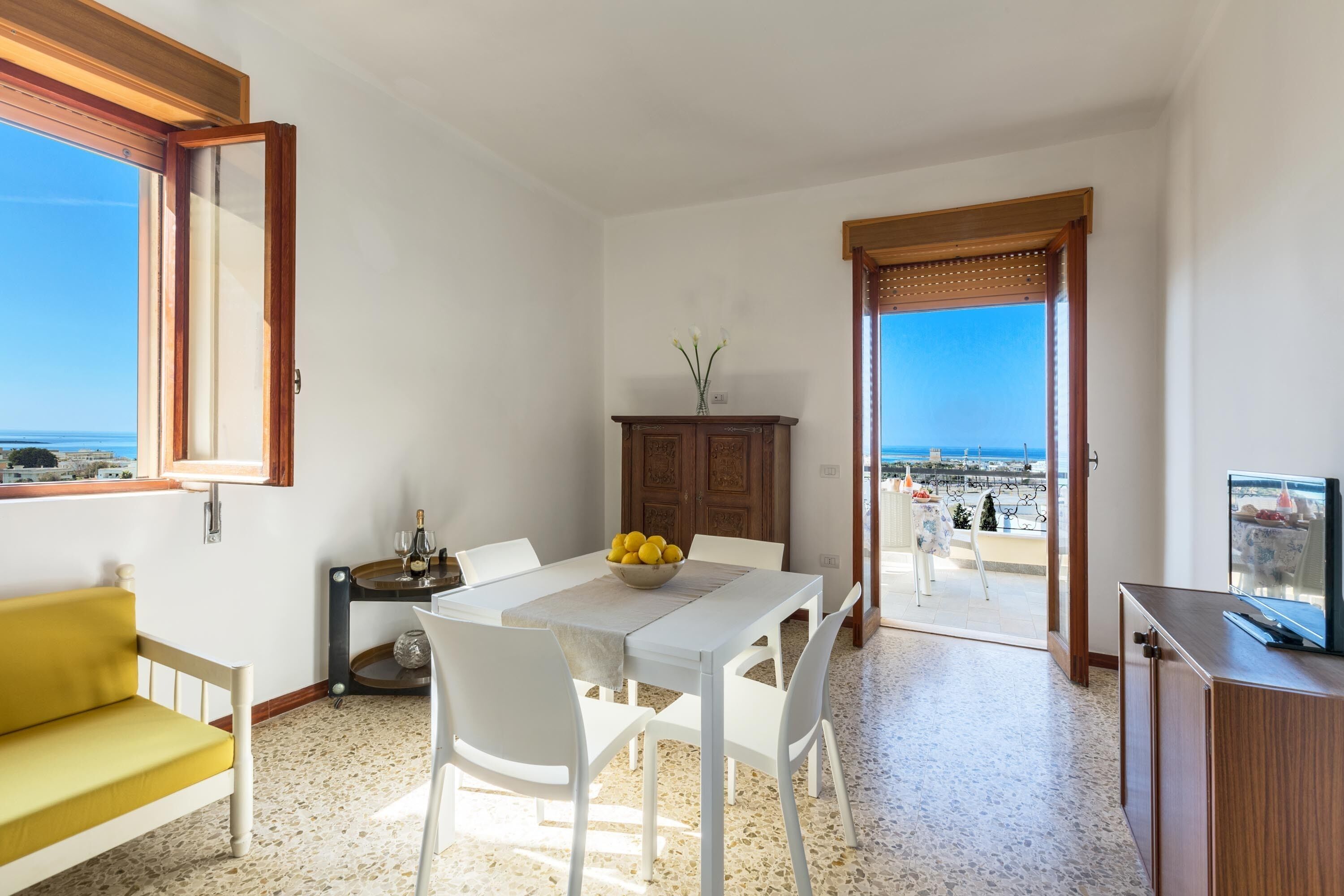 A dining area with a table, chairs, and a bowl of lemons. There is a sofa and a television on a cabinet.
