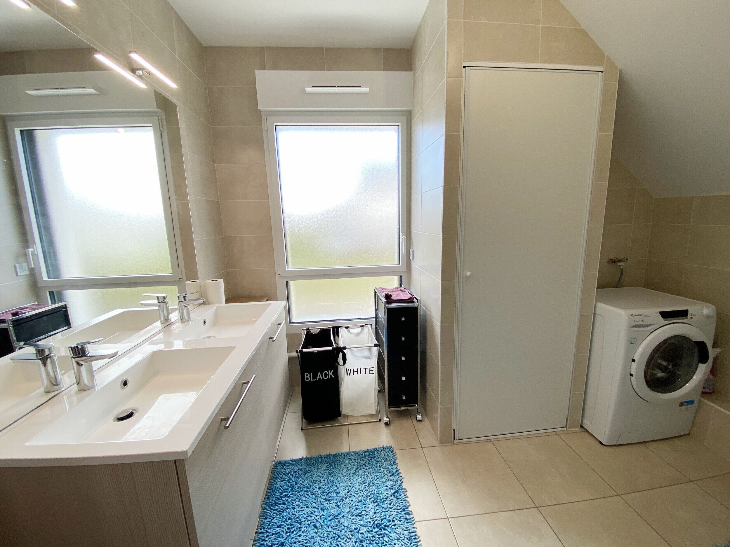 Une salle de bain moderne dotée d’un meuble-lavabo à deux vasques, d’un lave-linge et d’une grande fenêtre qui laisse entrer la lumière naturelle.