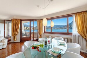 A dining area with a table set for two, a view of the sea and mountains, and a large window with curtains. - Comfort Holiday In Paradiso With Lake Lugano View (Paradiso)