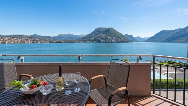 A balcony with a table set for two, overlooking a lake and mountains. - Comfort Holiday In Paradiso With Lake Lugano View (Paradiso)