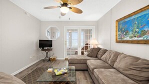 A living room with a beige sofa, a glass coffee table, a ceiling fan, a TV, and a painting of an underwater scene. - Nicely Renovated Bayfront Condo with Great Sunsets (Ocean City)