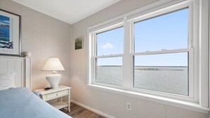 A bedroom with a large window, a bed, a nightstand, and a lamp. - Nicely Renovated Bayfront Condo with Great Sunsets (Ocean City)