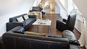 A modern living room with a sofa, armchair, and coffee table, a kitchen in the background, and a staircase leading to an upper level. - Herzlich Willkommen im Appartement Meier (List auf Sylt)