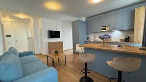 Een moderne woonkamer met een blauwe bank, een klein bijzettafeltje en twee barkrukken. Een flatscreen televisie is aan de muur bevestigd, en de keuken heeft een houten aanrechtblad en blauwe kastjes.